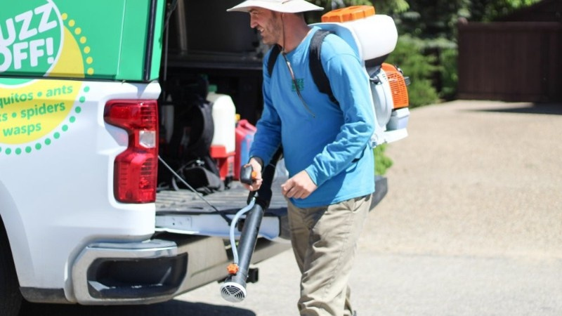 A pest control technician wearing a sun hat and a blue long-sleeve shirt, carrying a backpack sprayer and standing next to a Buzz Boss service truck
